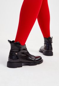 Botines de charol negro con parte trasera texturizada, cremallera lateral y suela gruesa. Llevados con medias rojas vibrantes, destacando el contraste.