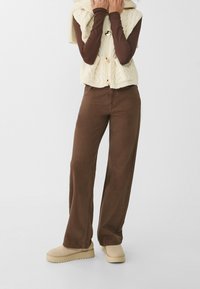 Vrouw in een bruine wijde broek, beige instappers, een bruine langemouwen top en een crèmekleurige kabelgebreide vest met grote knopen.