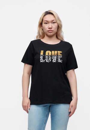 Mladá osoba so svetlými blond vlasmi má na sebe čierne tričko s textom v zlatej a bielej farbe "LOVE" a svetlomodré rifle, stojaca pred jednofarebným pozadím.