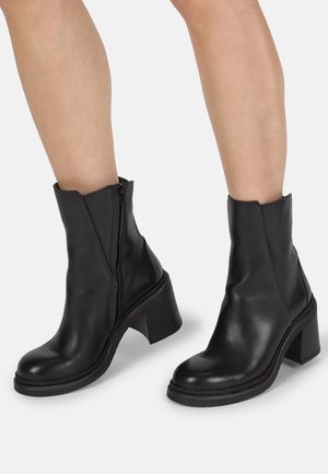 Bottines en cuir noir avec fermeture éclair latérale et talons blocs épais portées sur des jambes nues sur un fond clair uni.