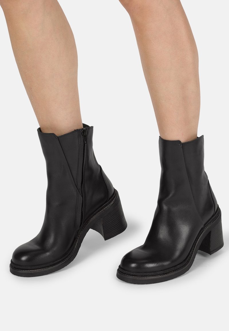 Bottines en cuir noir avec fermeture éclair latérale et talons blocs épais portées sur des jambes nues sur un fond clair uni.