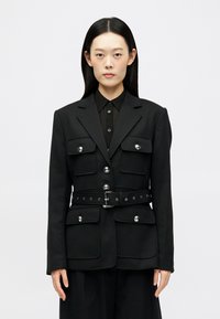 Musta muotopuku takki, jossa on pehmustetut olkapäät, hopeiset napit ja isot taskut. Siinä on vyötärölle tuleva vöitä ja strukturoitu muotoilu.