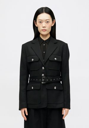 Musta muotopuku takki, jossa on pehmustetut olkapäät, hopeiset napit ja isot taskut. Siinä on vyötärölle tuleva vöitä ja strukturoitu muotoilu.