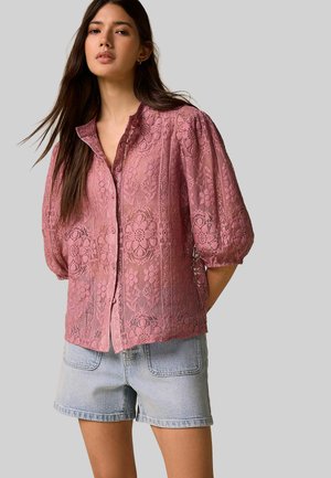 Jeune femme portant un chemisier en dentelle rose transparente avec des motifs floraux et des manches bouffantes, associé à un short en jean clair, debout devant un fond gris.