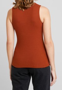 Kvinna klädd i en ärmlös, rostfärgad ribbad topp och svarta jeans, sedd bakifrån mot en neutral bakgrund.