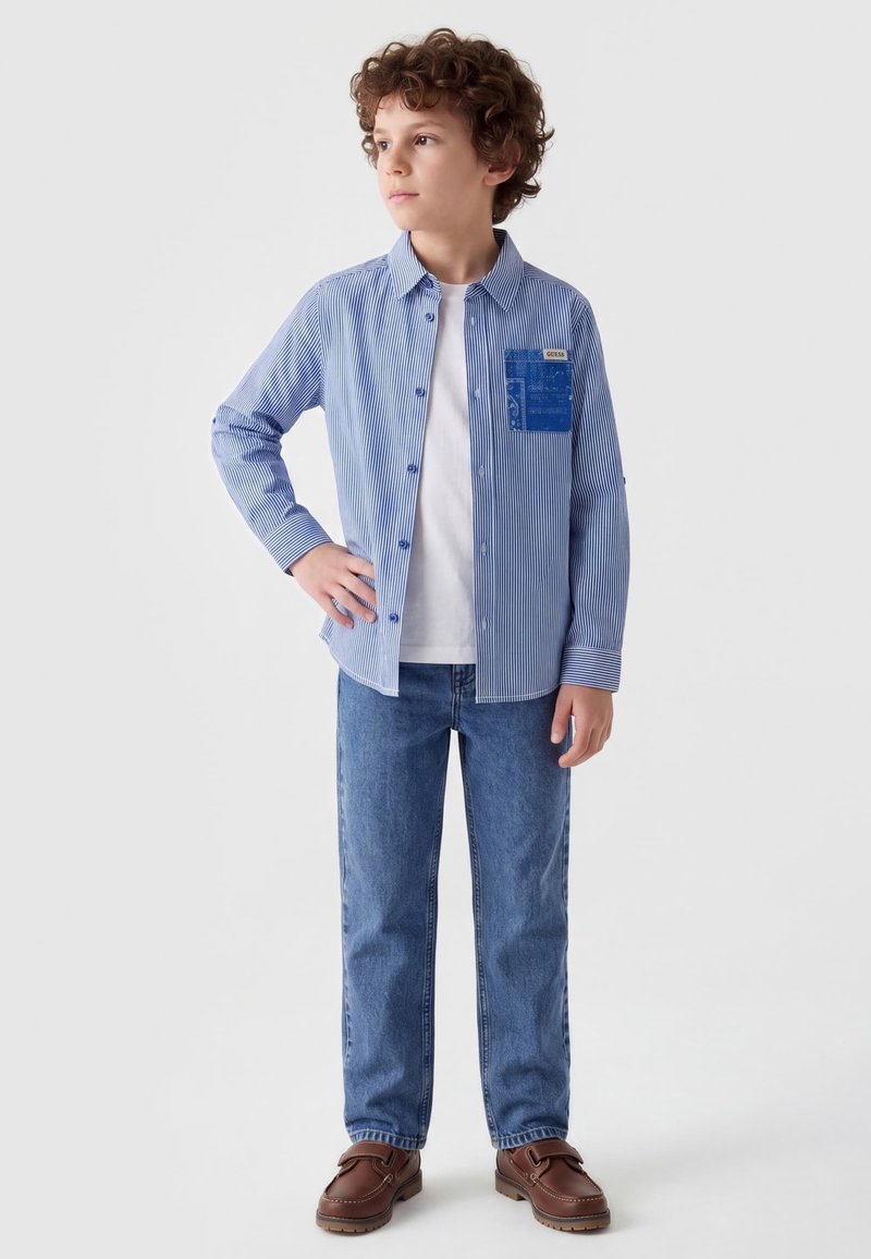 Garçon debout, main sur la hanche, portant une chemise rayée bleue par-dessus un t-shirt blanc, jean bleu et chaussures marron, regardant sur le côté sur fond blanc.