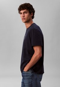 Tmavomodré bavlnené tričko s krátkymi rukávmi, okrúhlym výstrihom a zaobleným lemom; na prednej strane s grafickým dizajnom; nosené s modrými džínsami.