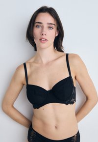 Sutiã de renda preta com um design estruturado, suporte com aro e alças ajustáveis. Os detalhes incluem um delicado acabamento em renda e um tecido texturizado.