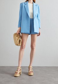 Blazer azzurro chiaro e pantaloni coordinati con una maglietta bianca texture. Gli accessori includono una clutch intrecciata dorata e sandali platform beige.