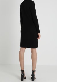 Robe en maille côtelée noire à manches longues, longueur mi-mollet, col montant et fentes sur les côtés. Portée avec des bottines noires à talons hauts.