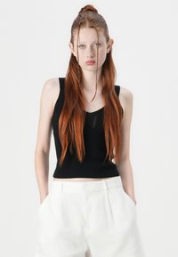 Top negru din tricot, fără mâneci, cu un design texturat, asortat cu pantaloni scurți albi din in, cu talie înaltă. Păr lung și drept, roșu.