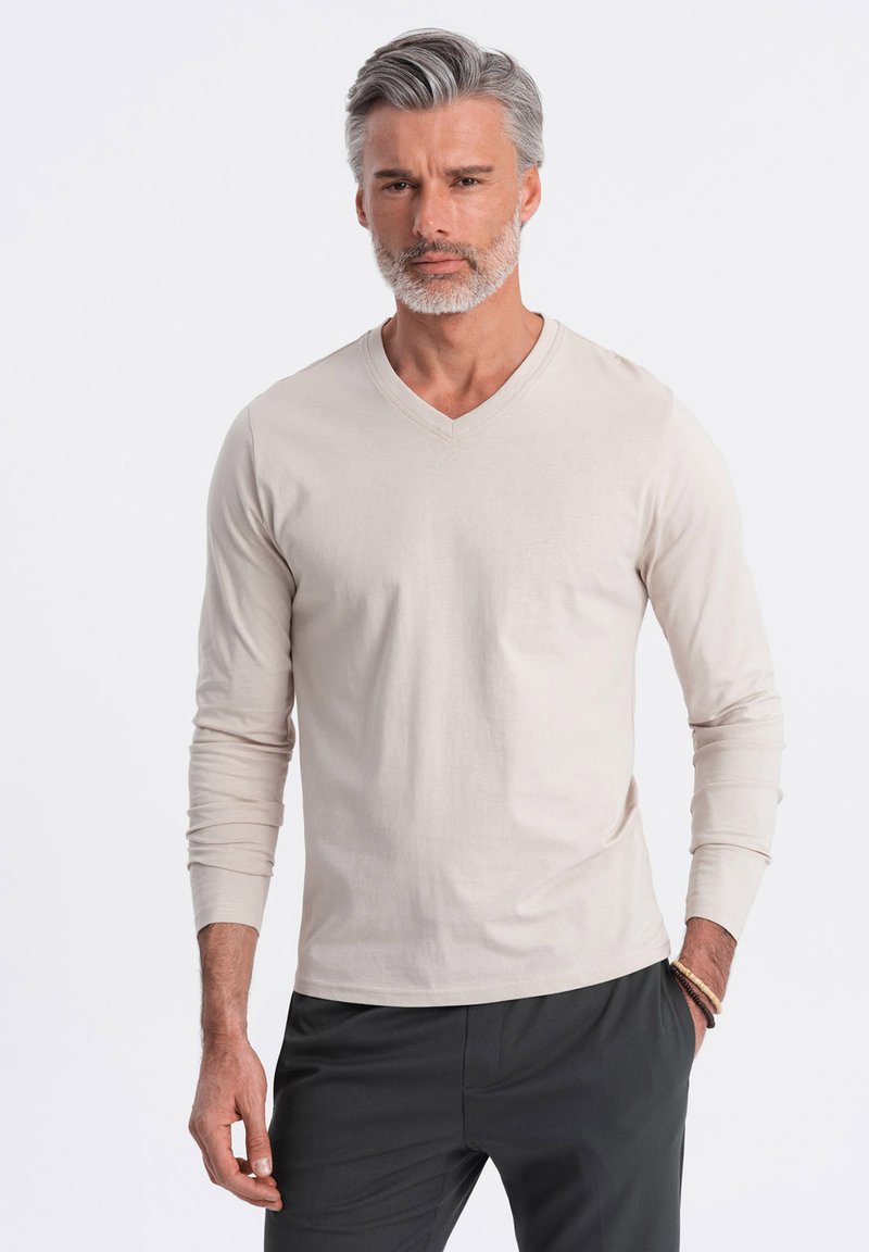 Langärmliges, V-Ausschnitt-T-Shirt aus hellbeigem Stoff mit glatter Textur. Kombination mit dunklen Hosen, hat enganliegende Ärmel und einen klaren Schnitt.