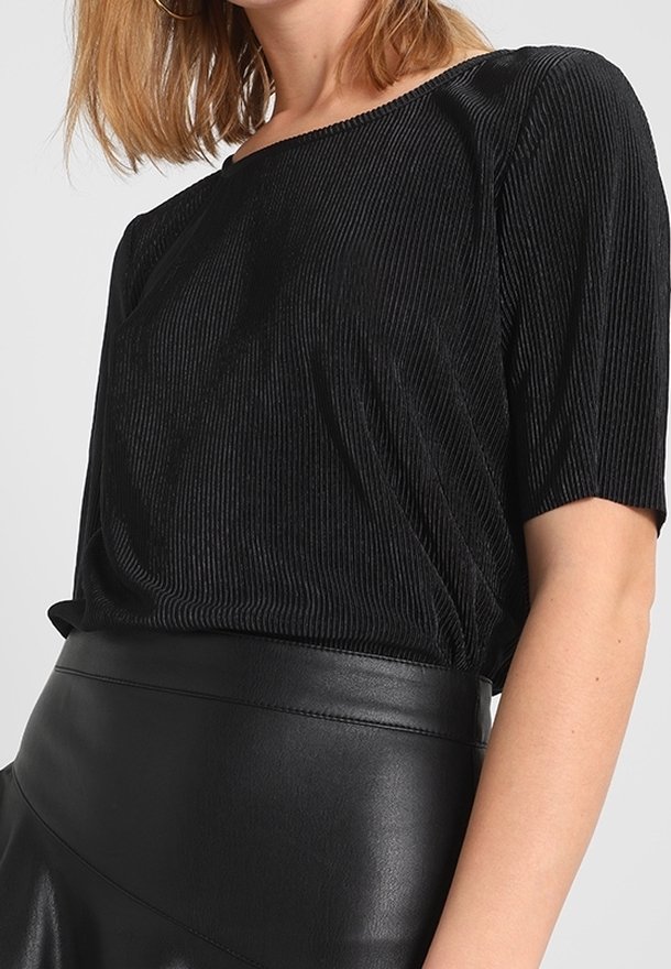 Femme portant un haut noir côtelé à manches courtes rentré dans une jupe en cuir noire taille haute sur un fond uni.