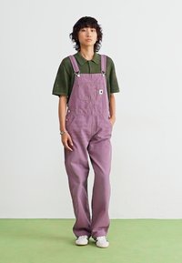 Salopette di denim viola con spalline regolabili e tasca frontale, abbinata a una camicia verde con colletto, su uno sfondo neutro.