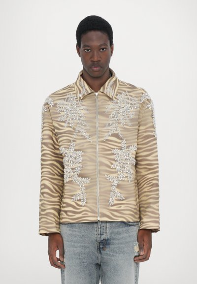 Beige jakke med zebra-stribet mønster, prydet med sølvfarvede blomsterpailletten, med krave og lynlås foran.
