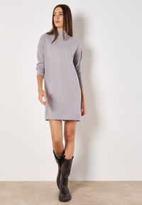 Robe tricotée côtelée grise avec un col haut et des manches longues, associée à des bottes en cuir noires hautes. Texture lisse, silhouette décontractée.