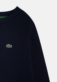 Tmavomodrý sveter vyrobený z pleteného materiálu, s rebrovaným golierom a malým zeleným vyšívaným logom krokodíla na hrudi.