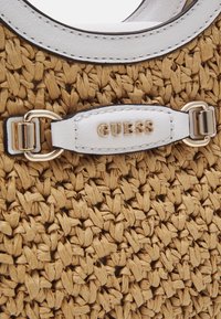 Détail d'un sac en paille tissée avec garniture en cuir blanc, ferrures dorées et logo « GUESS » sur une lanière en cuir blanc.