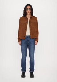 Veste en daim marron avec deux poches poitrine, portée sur une chemise beige, associée à un jean bleu et des chaussures noires, présentée sur un fond blanc.