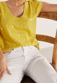 Žluté tričko s hlubokým výstřihem do V a krátkými rukávy, zdobené metalickými vzory mušlí. V kombinaci s bílými džínami.
