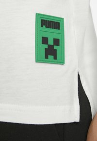 Tricou alb din bumbac, cu un patch verde din cauciuc al brandului Puma, având un design cu fața neagră a Creeper-ului, fante pe lateral și margine neagră la tiv.