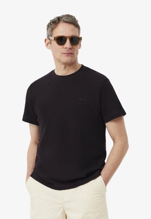 Hombre con gafas de sol carey, camisa negra de manga corta y pantalones beige claro con las manos en los bolsillos, mirando ligeramente hacia la izquierda.