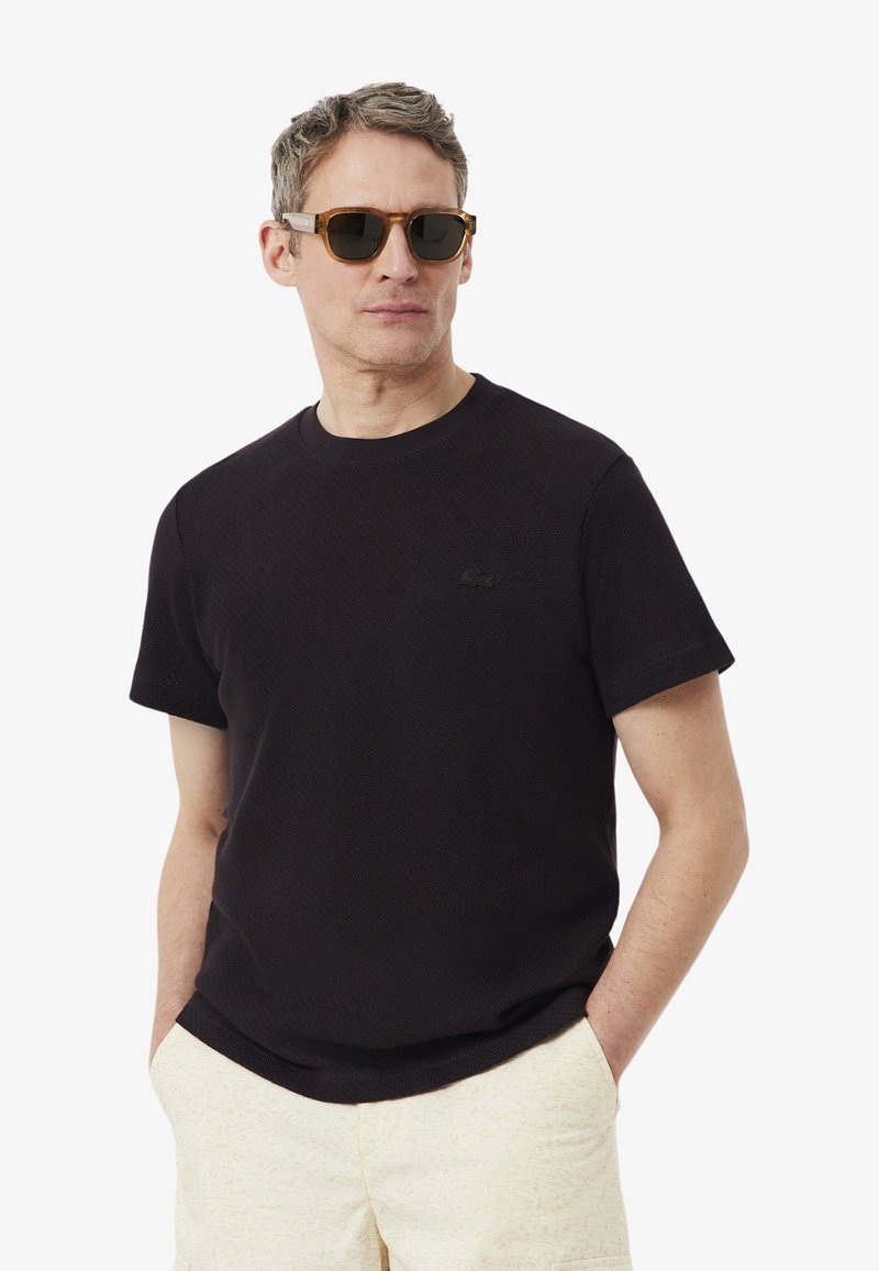 Hombre con gafas de sol carey, camisa negra de manga corta y pantalones beige claro con las manos en los bolsillos, mirando ligeramente hacia la izquierda.