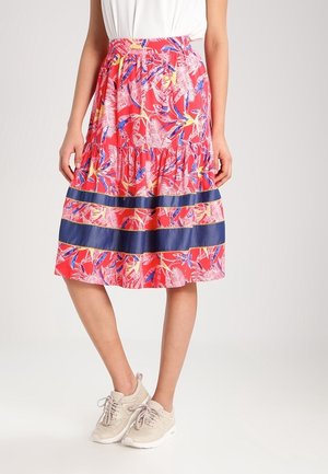 Femme portant une jupe midi rouge à fleurs avec des rayures horizontales bleues, associée à un haut blanc et des baskets beiges sur un fond uni.