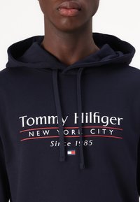 Felpa navy con logo frontale "Tommy Hilfiger", caratterizzata da dettagli rossi e cordini. Tessuto morbido, vestibilità casual.