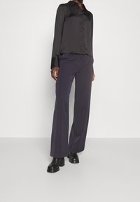Chemise noire en satin boutonnée associée à un pantalon sur mesure violet foncé. Le modèle porte des bottines noires à talons épais. Design simple et élégant.