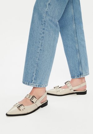 Persoon draagt lichtblauwe spijkerbroek en crèmekleurige slingback platte schoenen met spitse neus en zilveren gespen op een witte achtergrond.