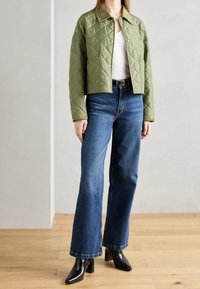 Grøn quiltet jakke med trykknapper, parret med blå buksere med vidde. Sorte spidse hæle fuldender outfittet. Trægulv i baggrunden.