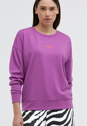 Frau trägt einen violetten Pullover mit dem Text "senses THE LABEL" und schwarz-weiß gestreifte Zebrahose, steht vor einem einfarbigen Hintergrund.