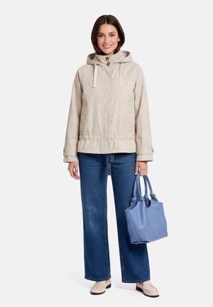 Woman wearing beige hooded jacket and blue jeans, holding a light blue handbag, standing and smiling against a white background.