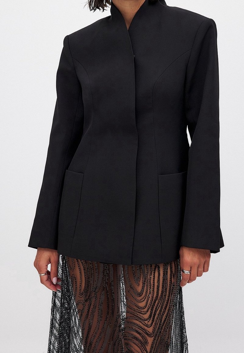 Femme portant un blazer noir ajusté sans boutons visibles et une jupe transparente avec des motifs noirs tourbillonnants.
