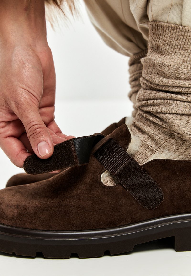 Scarpa in suede marrone scuro con chiusura a strappo nera, indossata con calzini beige a coste, su sfondo bianco.