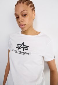 T-shirt blanc à col rond, arborant un logo « ALPHA INDUSTRIES » noir et un design graphique au centre. Texture en coton lisse.