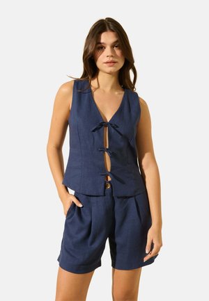 Top sans manches en lin bleu marine avec un décolleté en V profond et trois fermetures à nœud à l'avant, associé à un short plissé assorti.