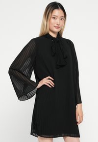 Μαύρο πλισέ φόρεμα με μακριές, πλατιά μανίκια και κορδόνι στον λαιμό. Φτιαγμένο από ελαφρύ ύφασμα, διαθέτει λεία υφή και διαφάνειες.