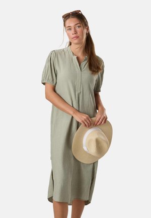 Kvinde i lysegrøn midi-kjole med korte ærmer holder en beige bredskygget hat med solbriller på hovedet.