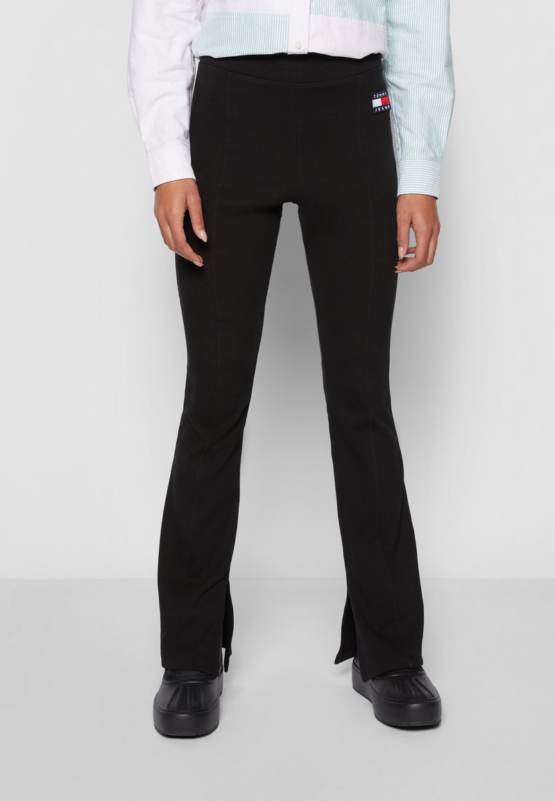 Personne portant un pantalon noir ajusté avec des fentes à l'avant et des bottes noires, associée à une chemise à manches longues boutonnée avec des panneaux bleu clair et blanc.