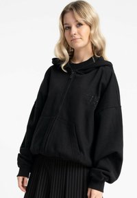 Czarna bluza rozpinana na zamek wykonana z miękkiego materiału, z dużym kapturem, opuszczonymi ramionami oraz małym geometrycznym wzorem na piersi.