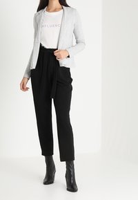 Šedé sako přes bílé tričko s fialovým nápisem "INFLUENCER", doplněné černými kalhotami s vysokým pasem a černými botami na podpatku.