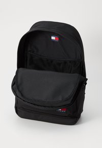 Sac à dos noir avec un compartiment principal comprenant une poche en mesh. Étiquette de logo remarquable sur le dessus. Tissu lisse avec une fermeture à glissière et un rabat.