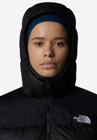 Fekete puffermellény magas gallérral, elöl cipzárral, és a The North Face logójával a mellkason. Simább textúrával és szigetelt kialakítással rendelkezik.