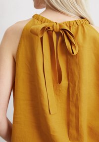 Mosterdgele mouwloze blouse met een geplooide halslijn en een strikdetail aan de achterkant, gemaakt van lichte stof.