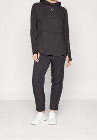 Svart atletisk huvtröja med framficka, långa ärmar och justerbara dragsnören, kombinerad med matchande svarta byxor och vita sneakers.