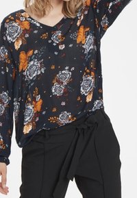 Blouse noire à fleurs présentant des fleurs orange et blanches, avec un décolleté en V et des manches longues. Assortie à un pantalon noir taille haute avec une ceinture.