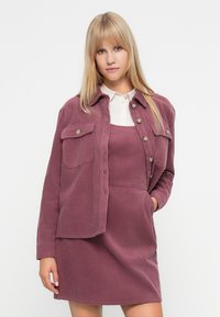 Σακάκι και φόρεμα από κωδωνί με ήπιο μωβ χρώμα. Το σακάκι έχει κουμπιά και δύο τσέπες στο στήθος, ενώ το φόρεμα διαθέτει εφαρμοστή γραμμή και τιράντες.