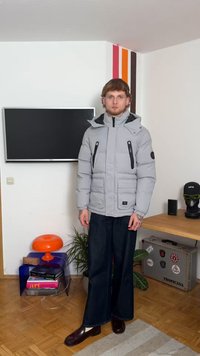 Ανοιχτό γκρι φουσκωτό μπουφάν με κουκούλα, που διαθέτει μαύρες τσέπες με φερμουάρ και ένα μπάλωμα. Συνδυάζεται με σκούρους φαρδιούς παντελονιού και γυαλιστερά παπούτσια.